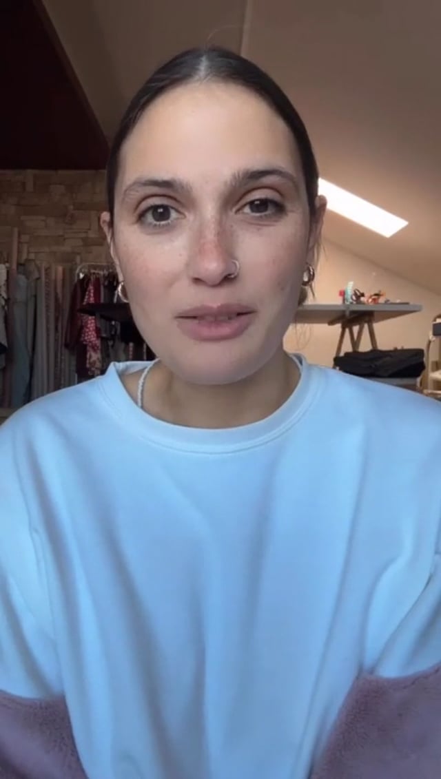 Fermo immagine del video: una donna con i capelli raccolti parla alla fotocamera in laboratorio, con gli stendini di tessuti alle spalle.