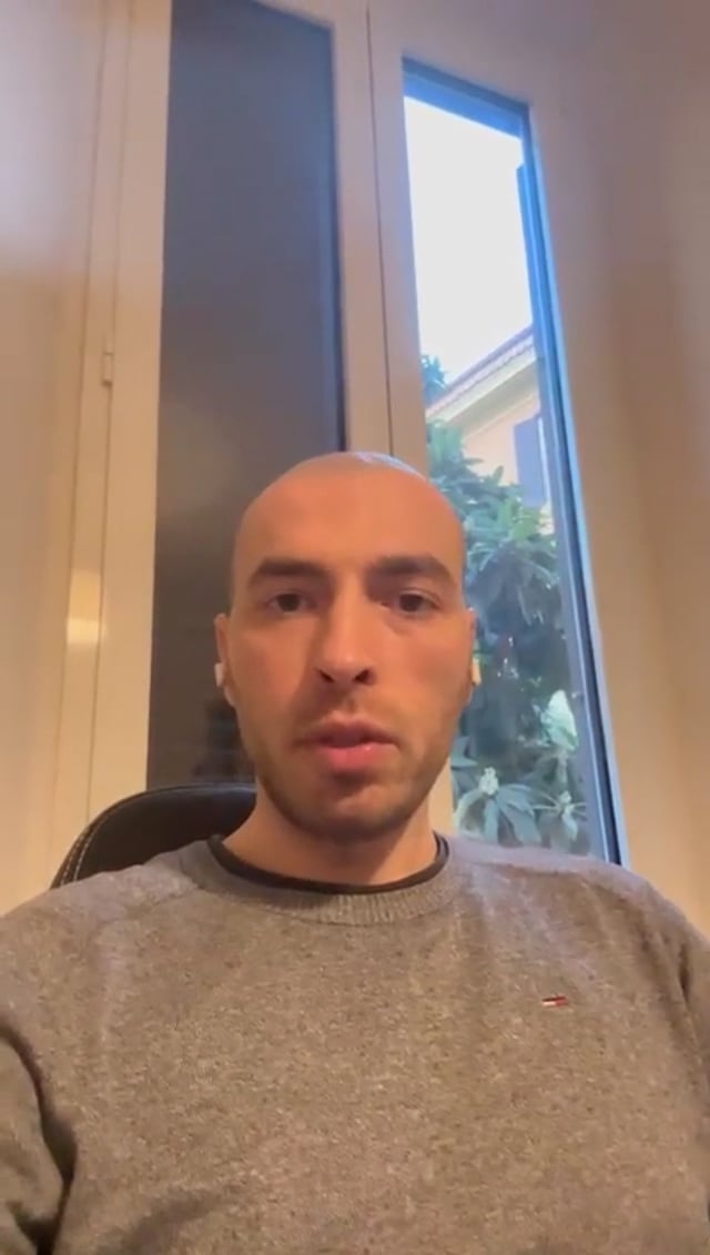 Fermo immagine del video: un uomo con gli auricolari, seduto alla scrivania davanti a una finestra, parla alla fotocamera del telefono.